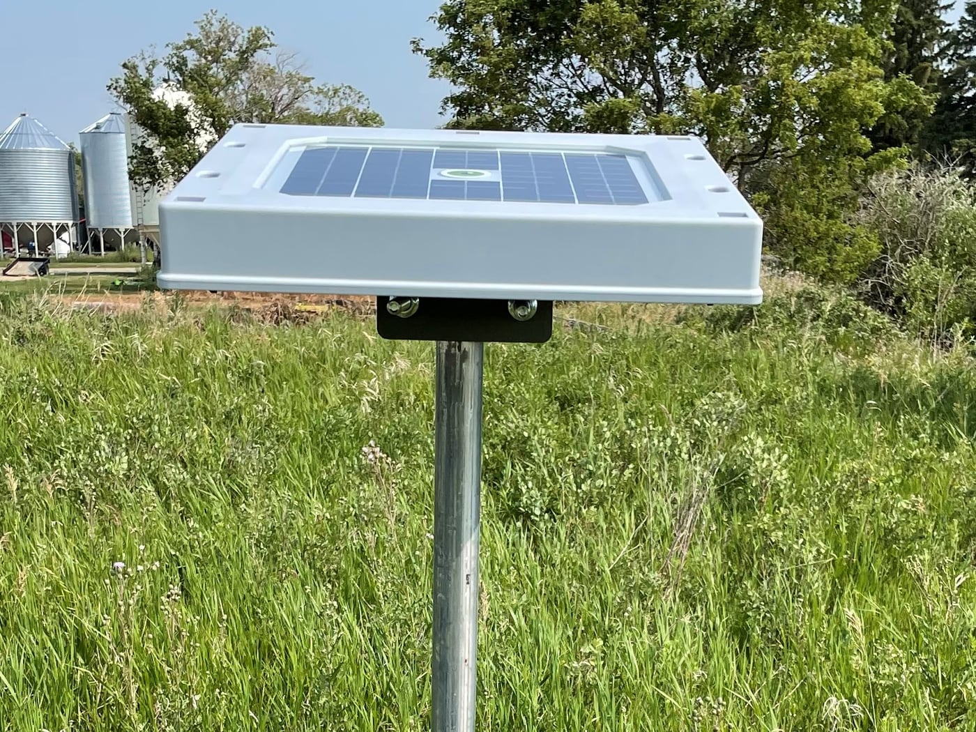 A pole-mounted Hailios Tessera sensor standing in a grassy field, grain bins and a farmyard behind it